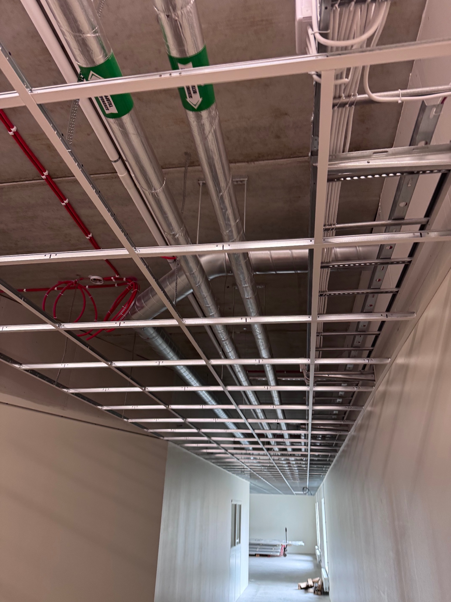 Sufit podwieszany w trakcie montażu w korytarzu biurowym. Widoczne instalacje: wentylacja z zielonym oznaczeniem, rury i kable w peszlach. Metalowa konstrukcja sufitu w perspektywie.