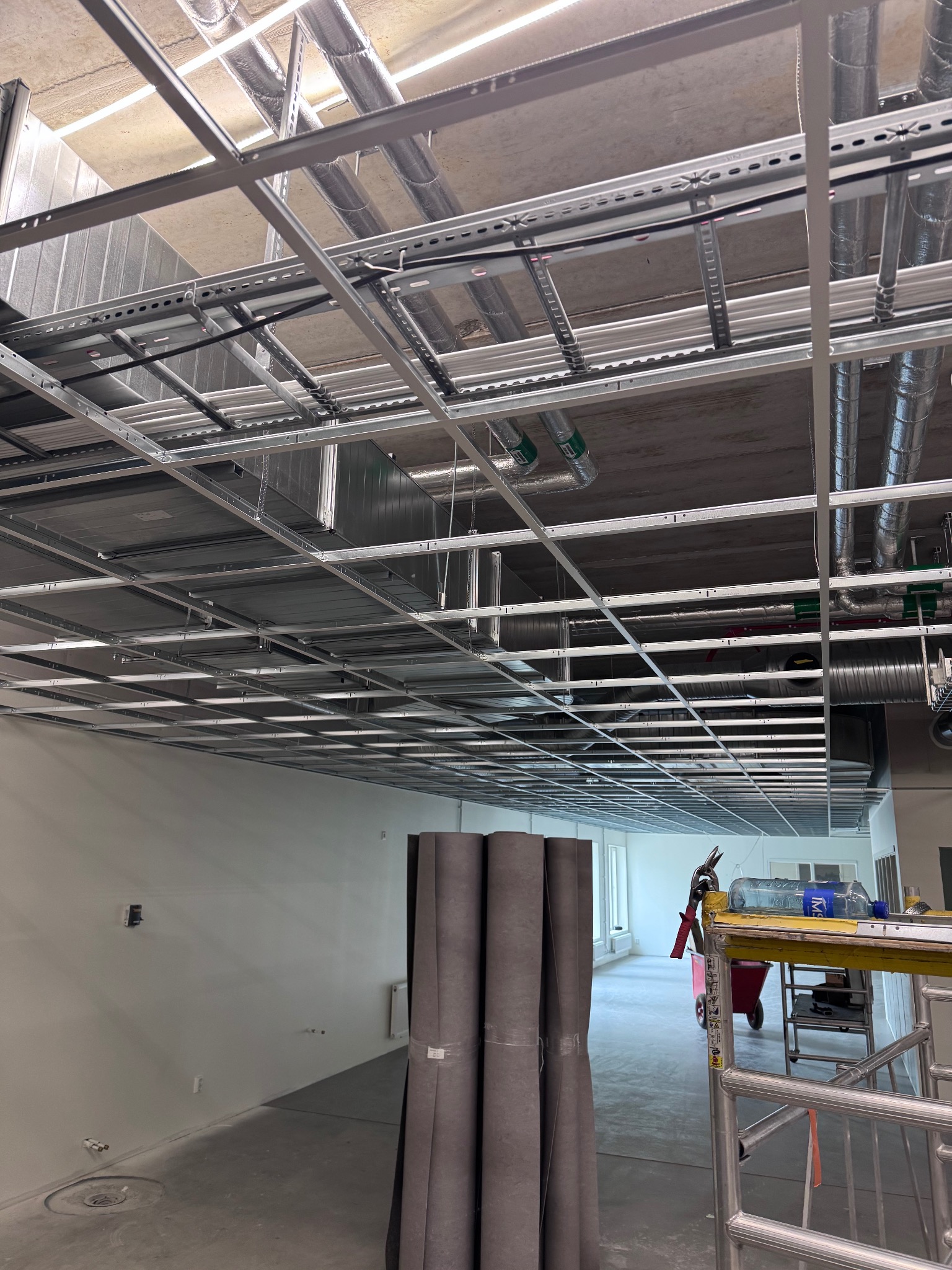 Metalowa konstrukcja sufitu podwieszanego w trakcie montażu, widoczne instalacje wentylacyjne i rolki materiału izolacyjnego w przestrzeni biurowej.