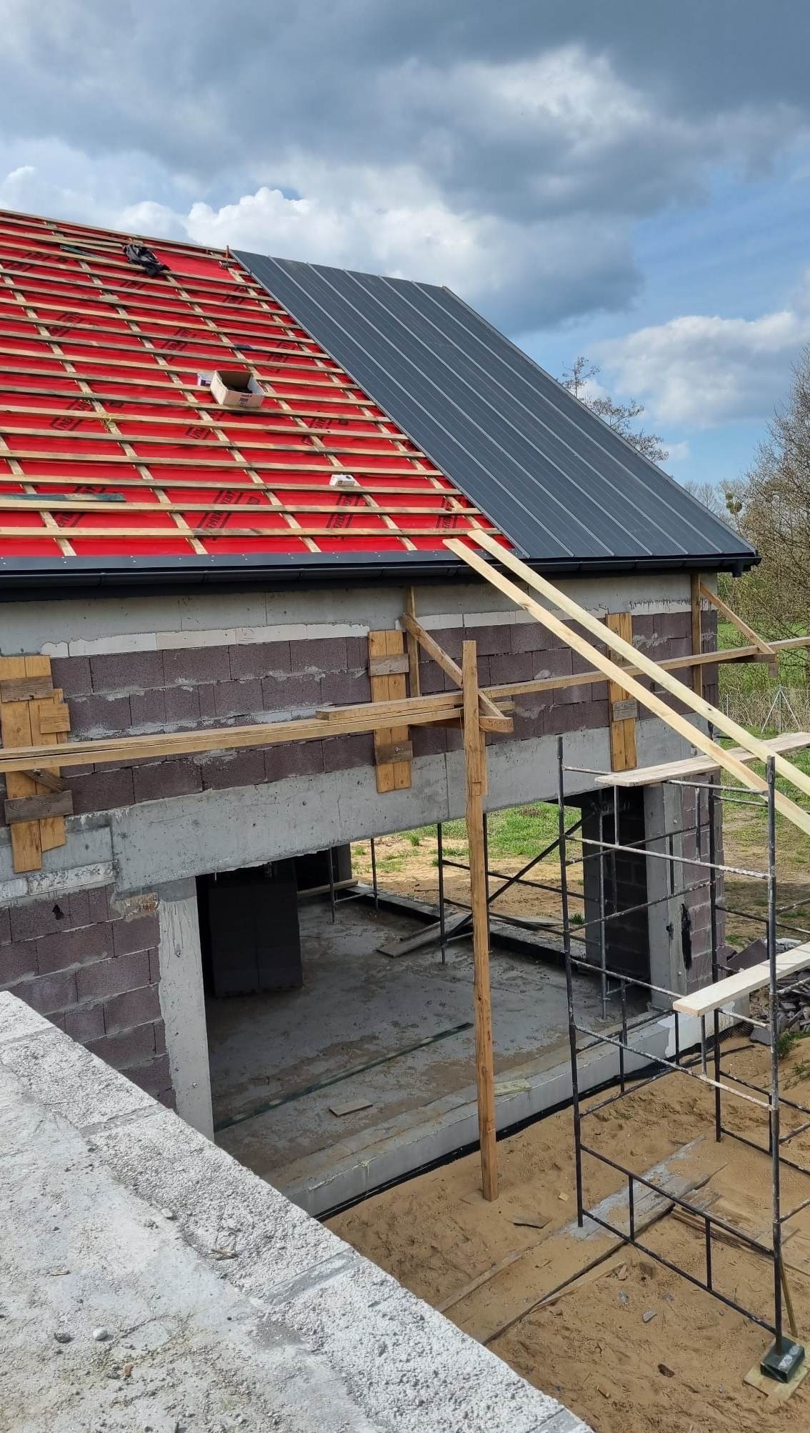 Budowa domu w toku: częściowo ułożona czerwona dachówka i szara blacha na dachu, widoczne rusztowania i surowy stan ścian budynku.