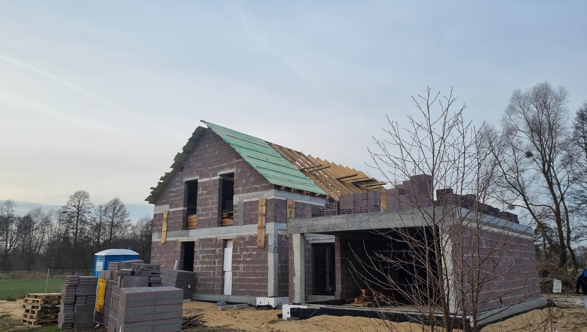 Surowy stan domu z pustaków: zielone deski na dachu, widoczne drewniane elementy konstrukcji, niezadaszony garaż, w tle drzewa i toaleta przenośna.