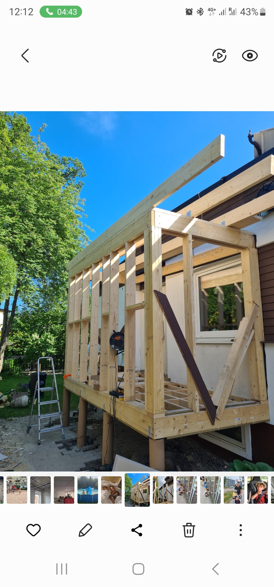 Drewniana konstrukcja dobudowy do domu w trakcie montażu. Widoczne słupy, rama, okno i narzędzia. Konstrukcja na zewnątrz budynku, zieleń w tle.