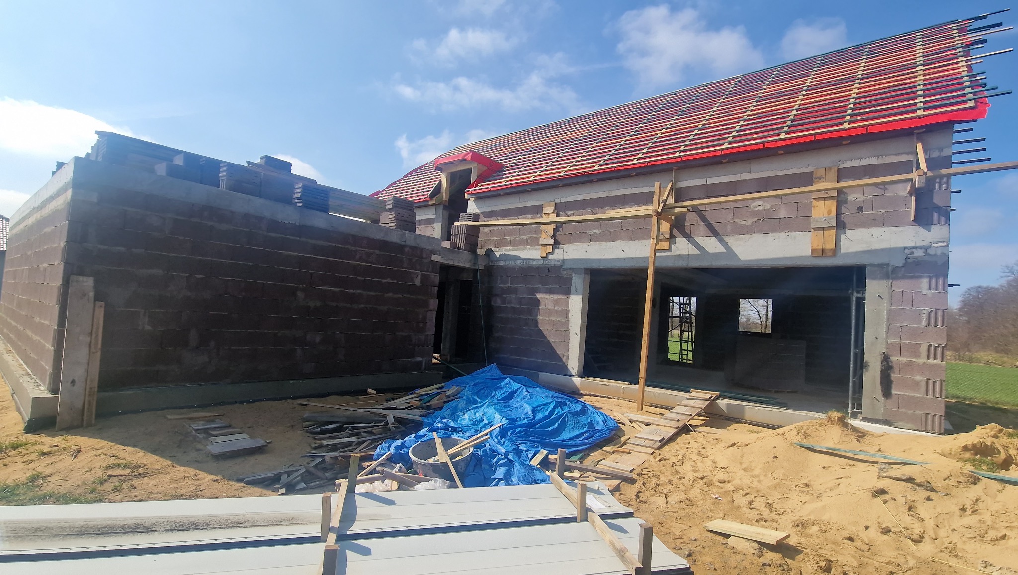 Budowa domu jednorodzinnego w trakcie realizacji: murowane ściany, więźba dachowa z łatami, niebieska plandeka, piasek. Widok na budynek z zewnątrz w słoneczny dzień.