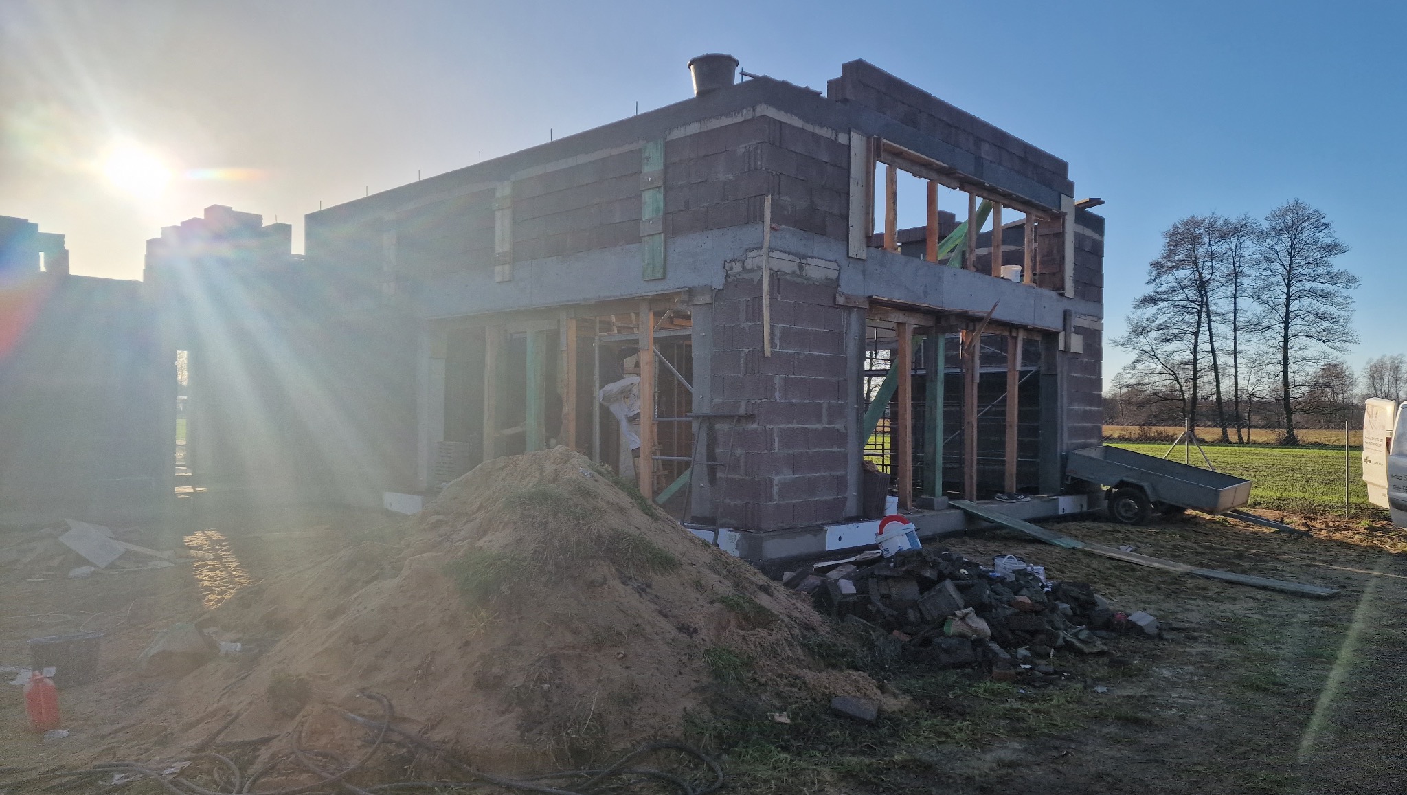 Budowa domu w trakcie realizacji: surowy stan z pustaków, widoczne drewniane szalunki okienne, w tle drzewa i zachodzące słońce. Na pierwszym planie sterta piasku.