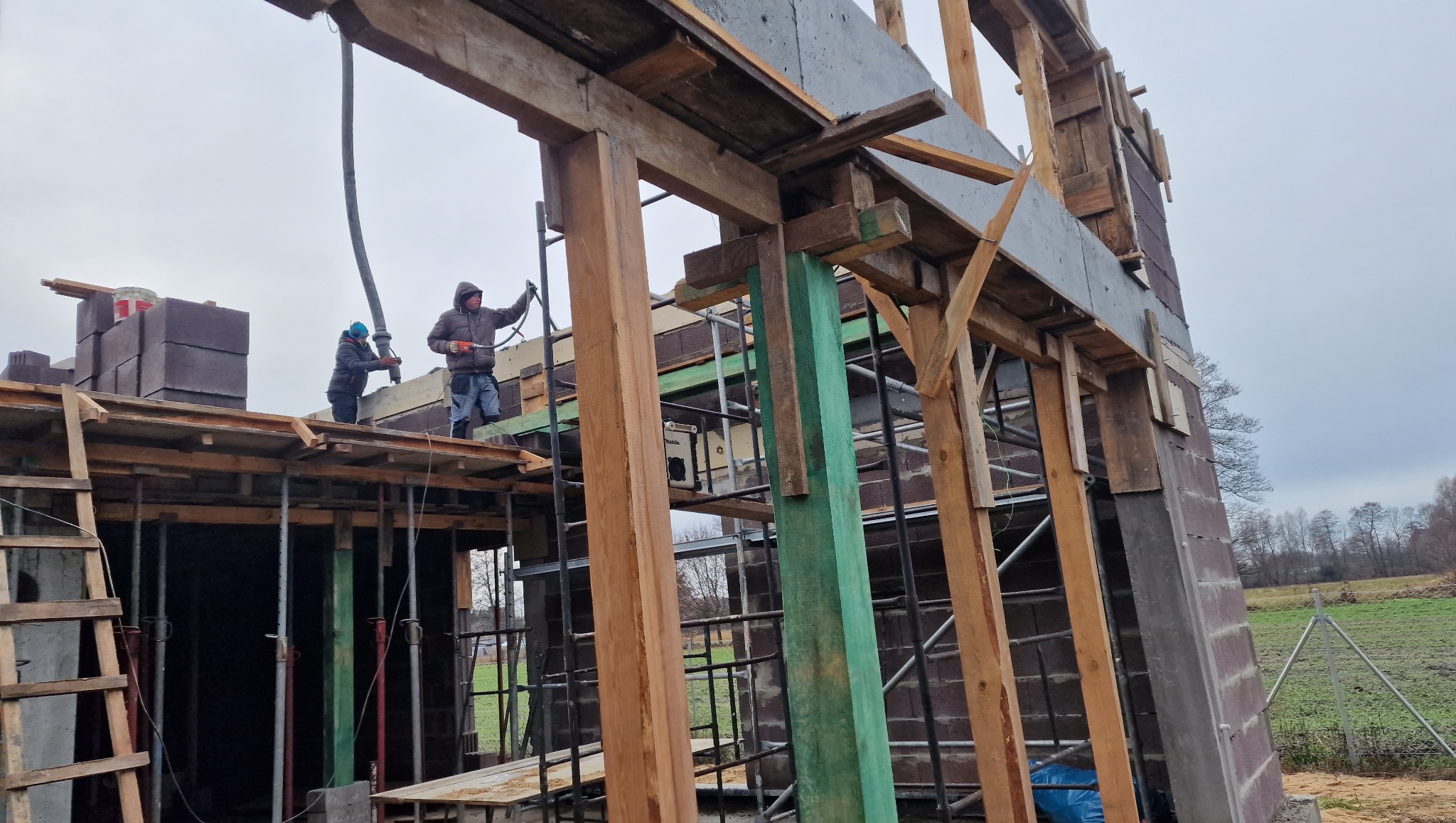 Budowa domu w toku: dwóch robotników na rusztowaniu pompuje beton do szalunków. Widoczne drewniane konstrukcje, bloczki i krajobraz wiejski w tle.