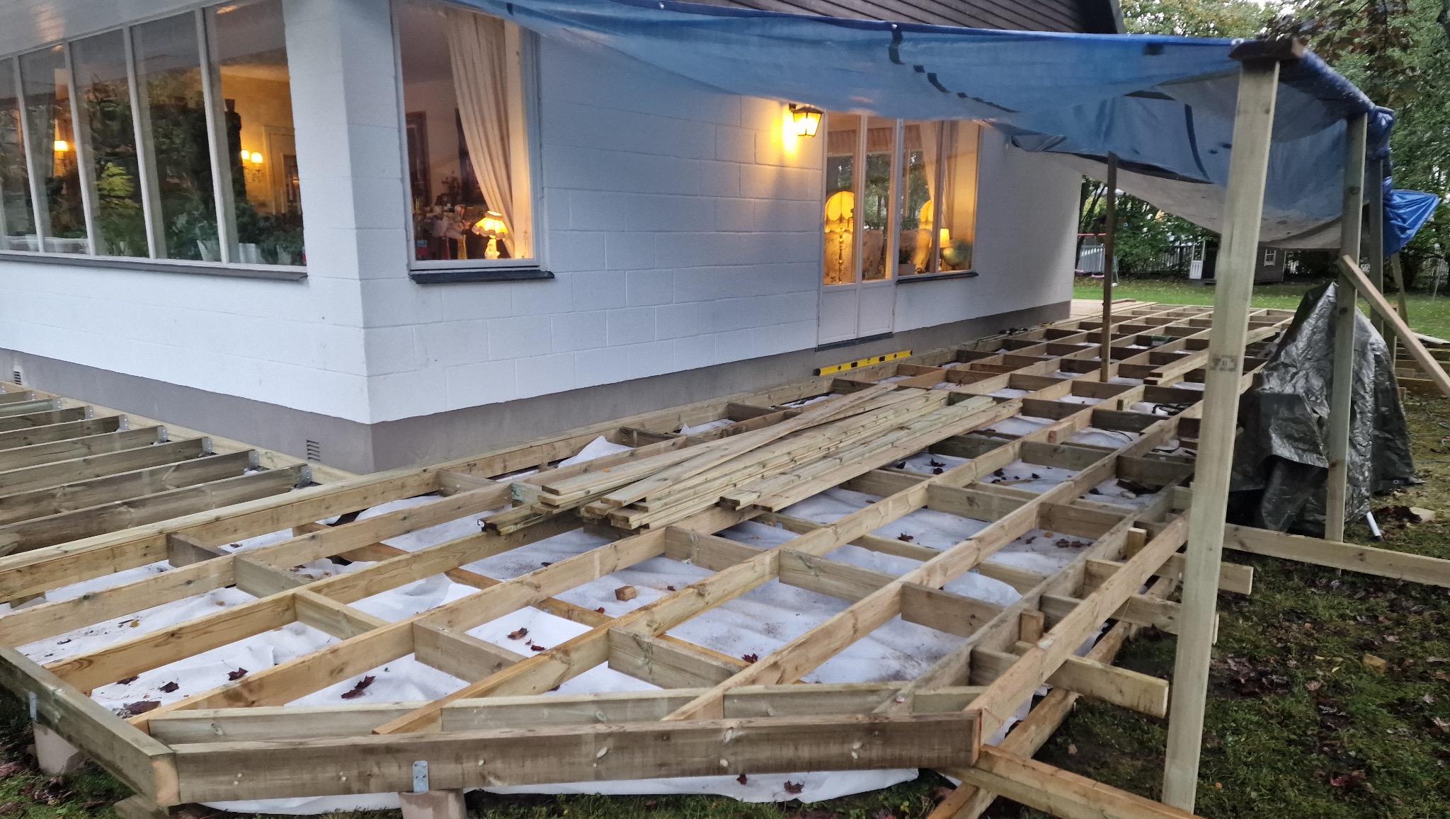 Konstrukcja drewniana tarasu przy białym domu z oknami, zabezpieczona folią. Widoczne elementy podtrzymujące i izolacja pod konstrukcją. Prace w toku, oświetlenie w oknach.