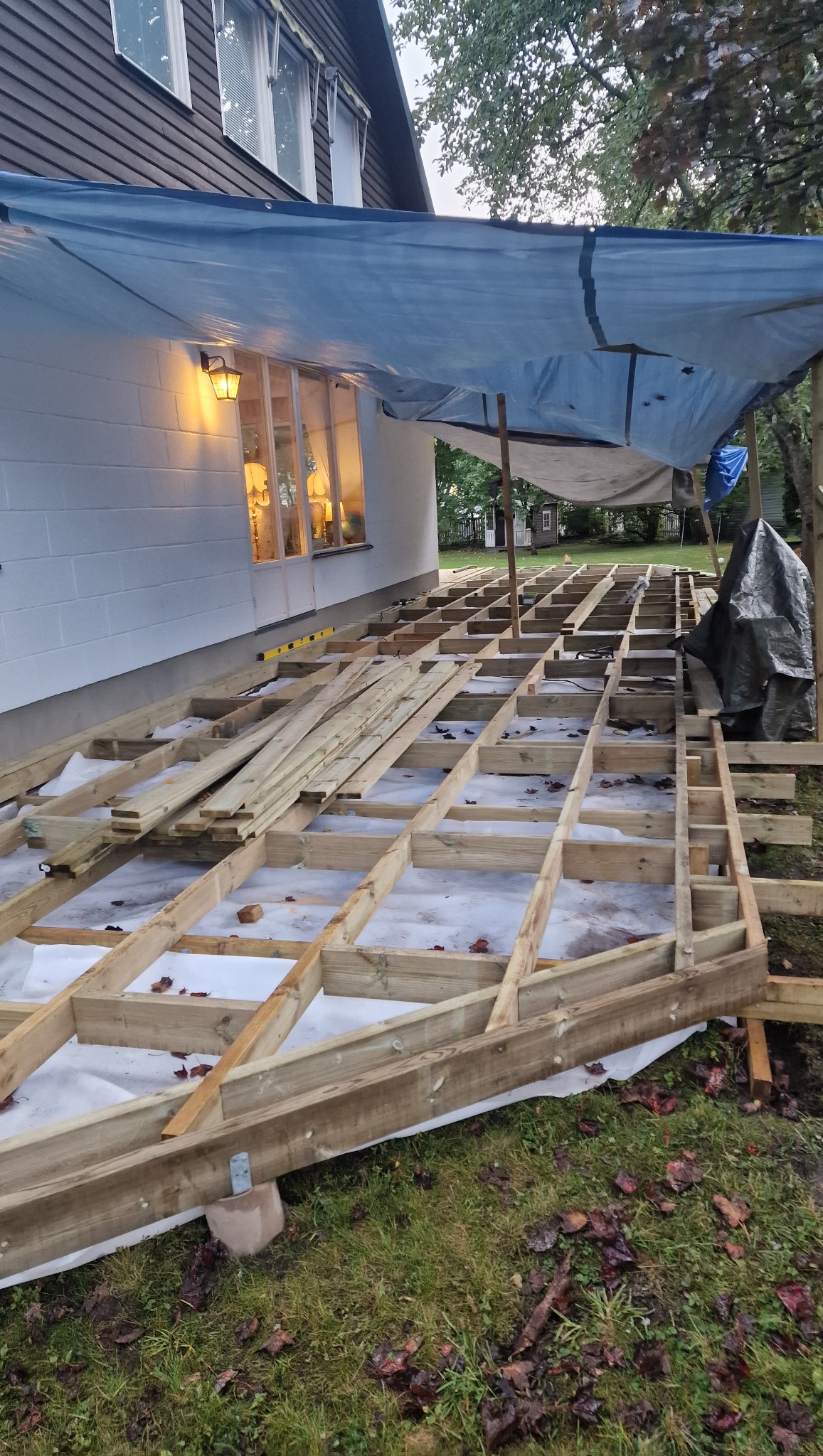 Konstrukcja drewnianego tarasu w trakcie budowy, widoczna rama z belek na białej membranie, zabezpieczona plandeką. Poziomica leży na belkach, okno z lampą w tle.