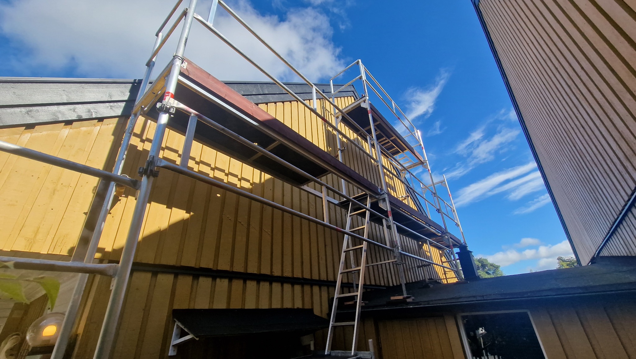 Żółta elewacja domu w trakcie prac na wysokościach, widoczna z dołu z fragmentem rusztowania i drabiny na tle błękitnego nieba.