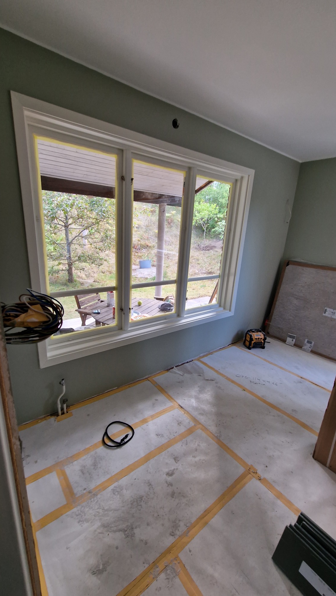 Pokój w trakcie prac wykończeniowych: okno zabezpieczone taśmą malarską, podłoga z zaznaczonym układem, widoczne narzędzia i materiały budowlane.