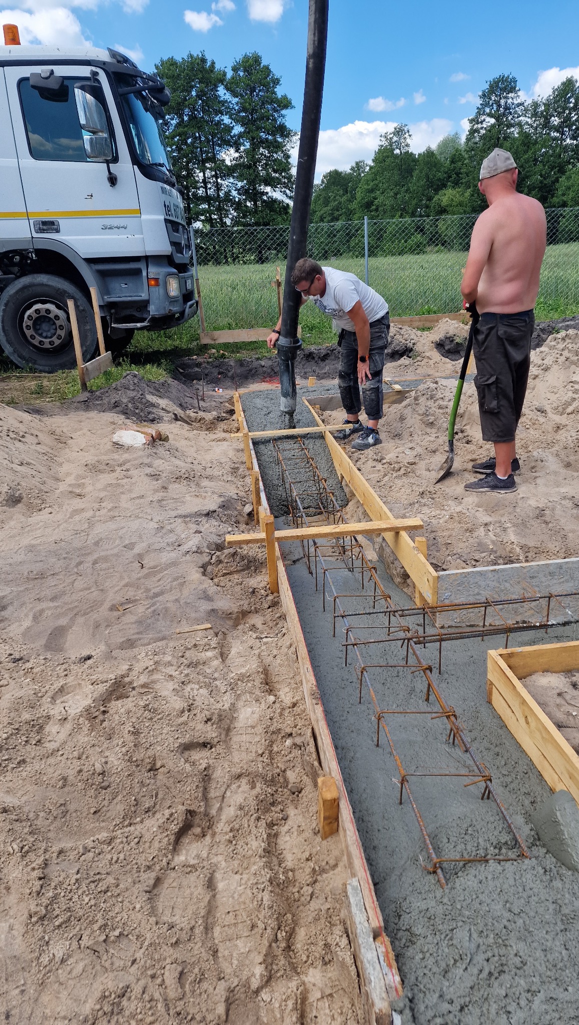 Wylewanie betonu do szalunku zbrojeniowego pod fundament budynku. Pracownicy obsługują pompę do betonu, widoczny samochód betoniarki i teren budowy.