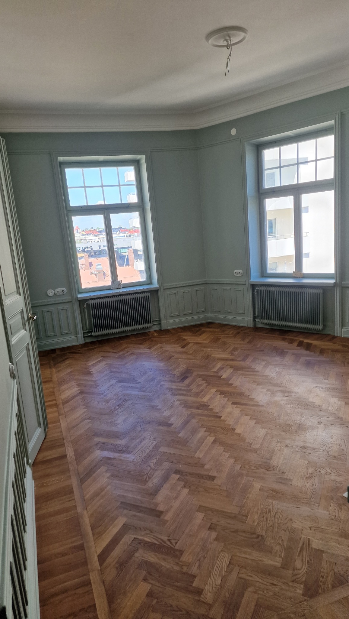 Pastelowy pokój z parkietem w jodełkę. Dwa okna z widokiem na miasto, grzejniki pod oknami, listwy przypodłogowe i sufitowe. Świeżo po remoncie, gotowy do umeblowania.