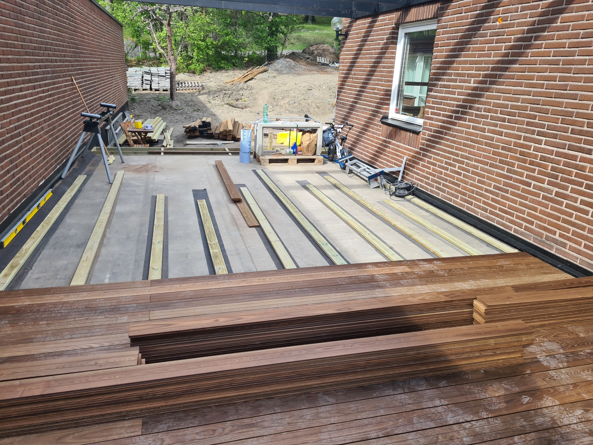 Montaż drewnianego tarasu: deski ułożone w stos, widoczne legary, narzędzia w tle. Ceglana ściana domu i zielone otoczenie dodają naturalnego charakteru.