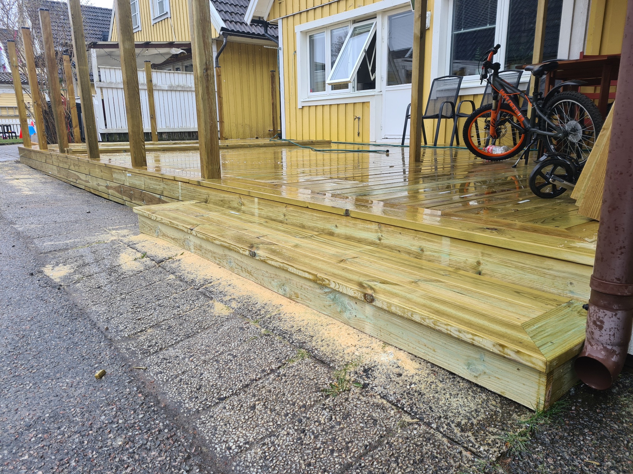 Nowy drewniany taras z elementami konstrukcyjnymi, świeżo po deszczu, z rowerem opartym o ścianę domu. Widoczne ślady obróbki drewna.
