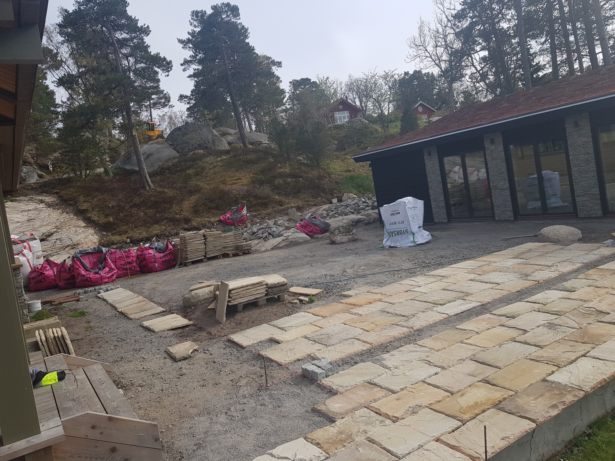 Układanie piaskowca na posesji domu w stylu skandynawskim. Widoczne worki z materiałami budowlanymi i narzędzia. Tło: skały, drzewa i dwa domy.