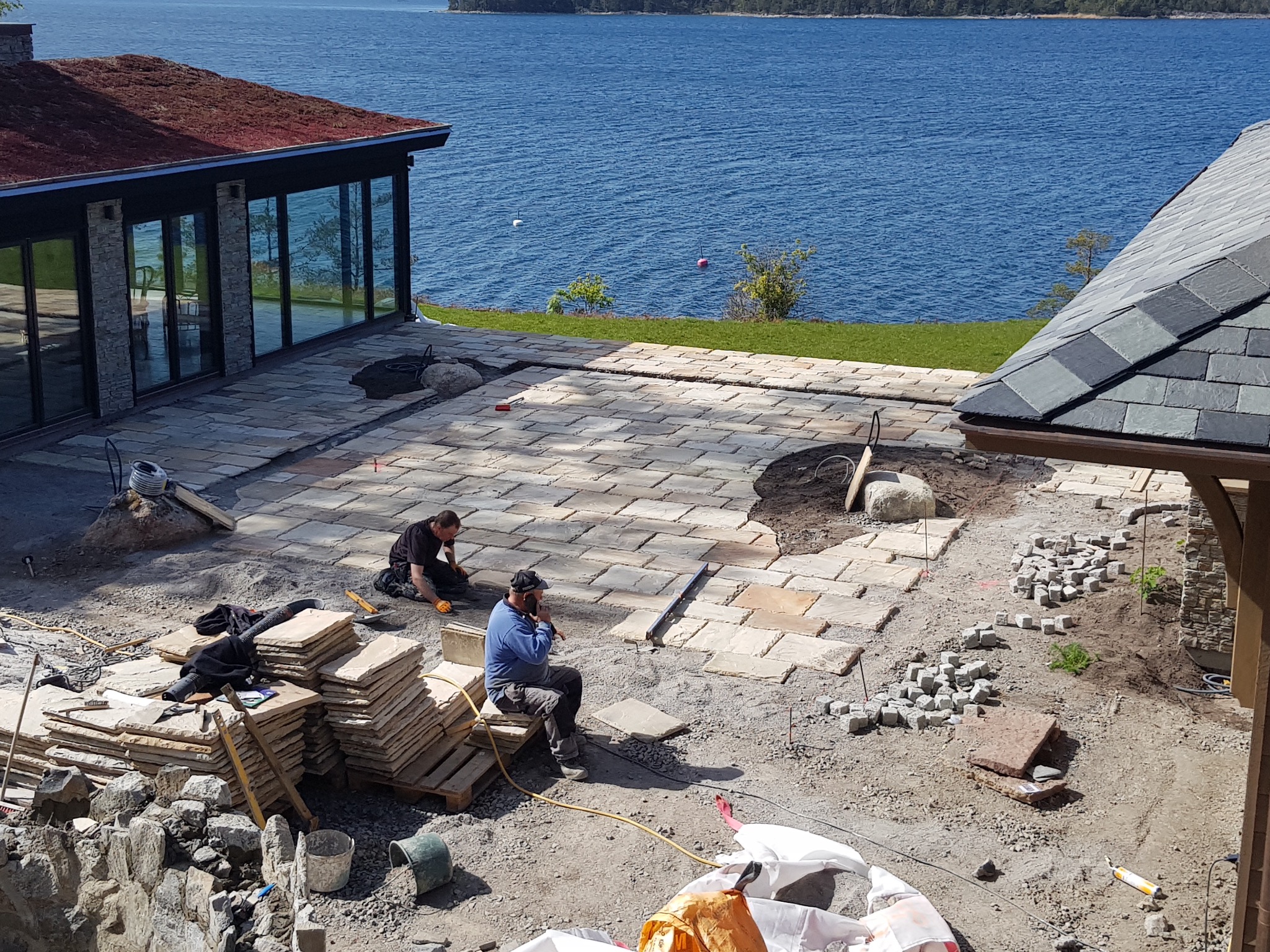 Układanie kamiennej posadzki na zewnątrz z widokiem na jezioro. Dwóch pracowników podczas pracy. Częściowo ułożona posadzka z kamienia naturalnego.