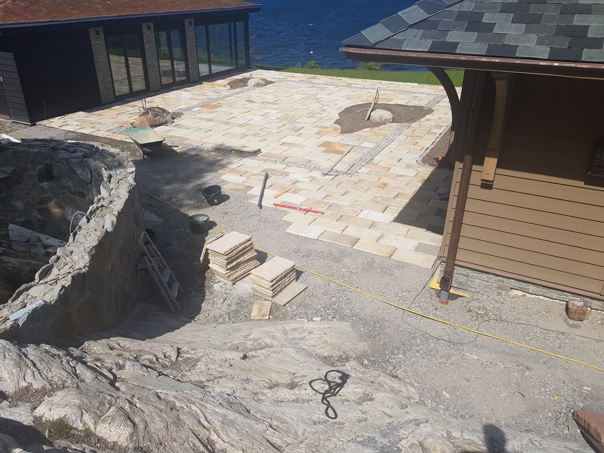 Układanie kamiennej nawierzchni tarasu z widokiem na jezioro. Częściowo ułożone płyty, narzędzia pracy i sterty kamieni na żwirowym podłożu. Widok z góry.