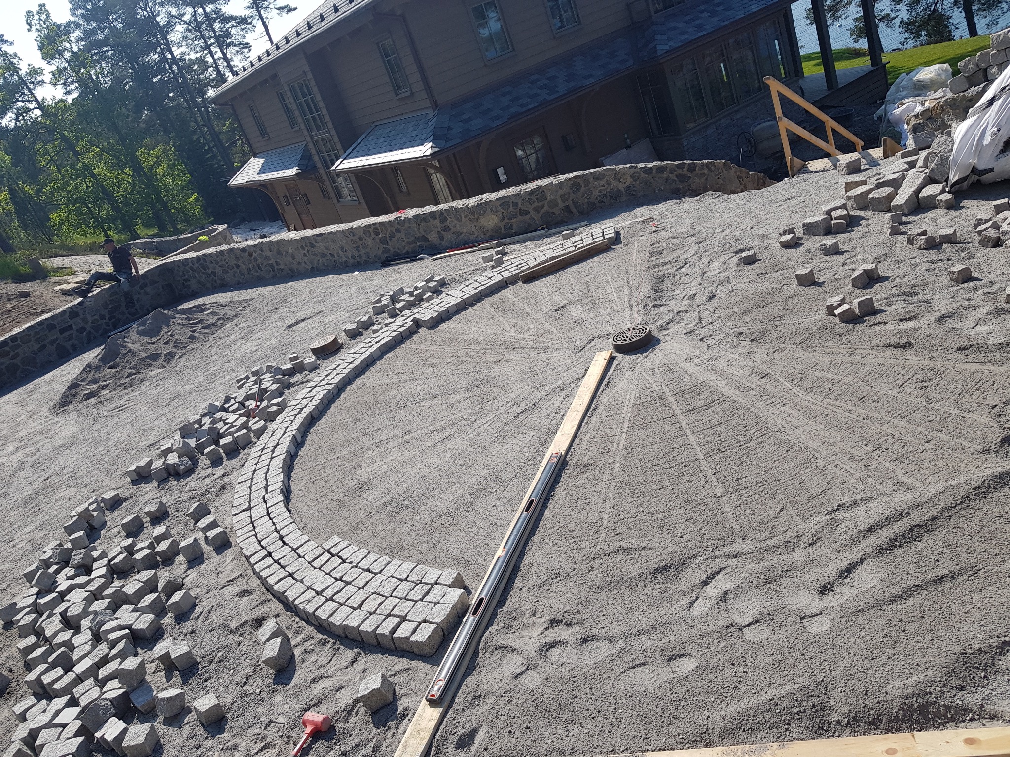 Układanie kostki brukowej na posesji przy domu z widokiem na jezioro. Część kostki ułożona w łuk, reszta w kupkach. Widoczne narzędzia i poziomica.