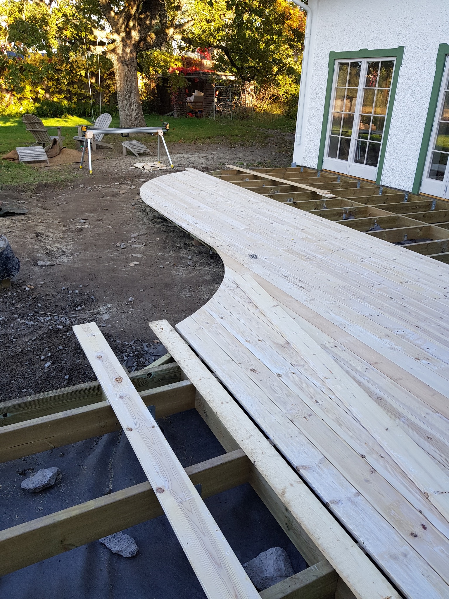 Budowa tarasu z desek o zaokrąglonym kształcie, widoczne elementy konstrukcyjne i deski, w tle budynek z białymi ścianami i zielonymi oknami.