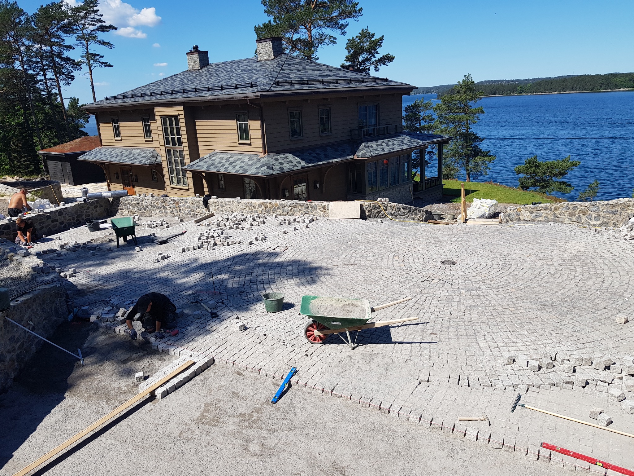 Układanie kostki brukowej na posesji w Grudziądzu. Widoczny fragment ułożonej nawierzchni, pracownicy i narzędzia. W tle dom i jezioro.