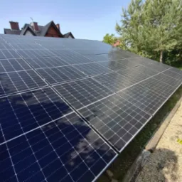 Panel słoneczny na dachu domu, widok z dołu, zieleń w tle. Instalacja fotowoltaiczna zamontowana na dachu, w tle drzewa i fragment domu z czerwoną dachówką.