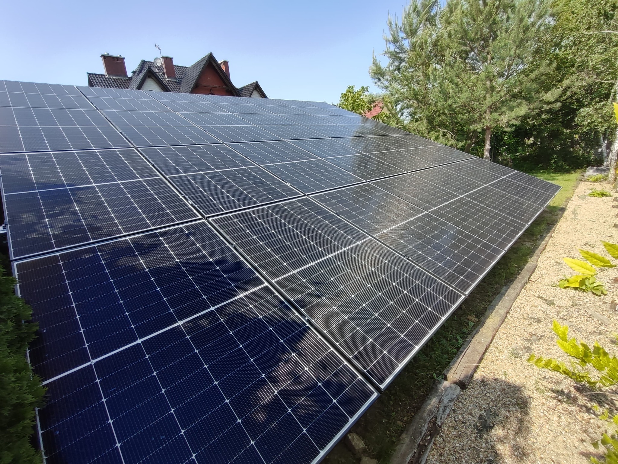 Panel słoneczny na dachu domu, widok z dołu, zieleń w tle. Instalacja fotowoltaiczna zamontowana na dachu, w tle drzewa i fragment domu z czerwoną dachówką.