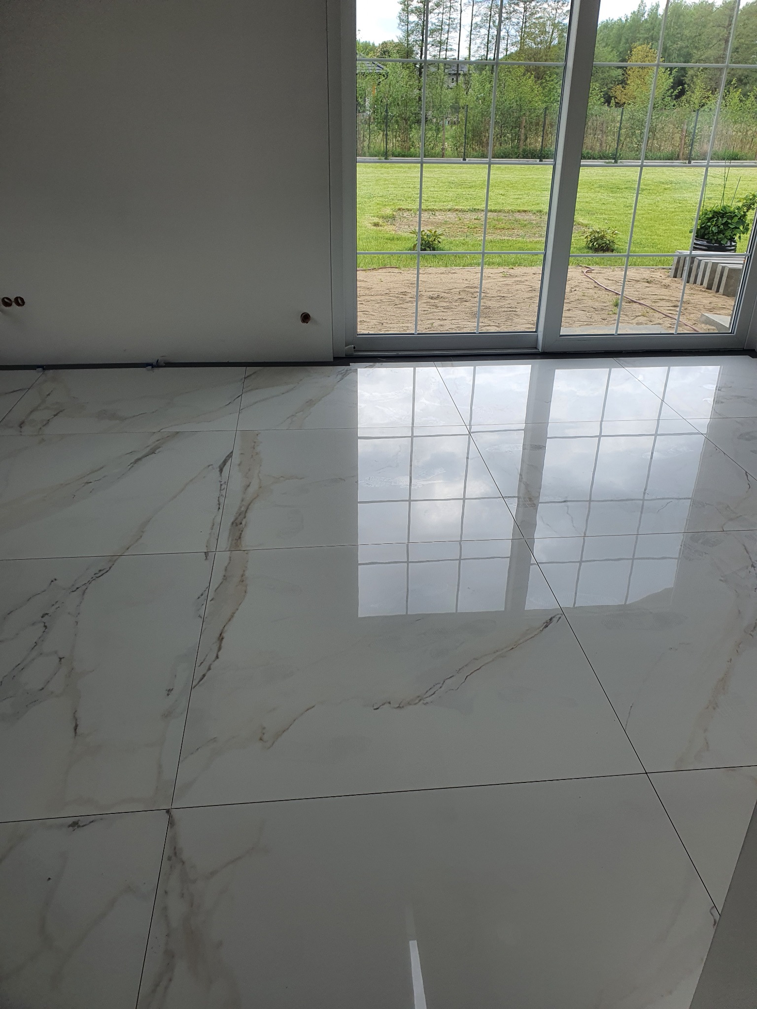 Błyszcząca, biała podłoga z marmurowym wzorem, ułożona z dużych kwadratowych płytek. Widok na ogród przez przeszklone drzwi balkonowe.