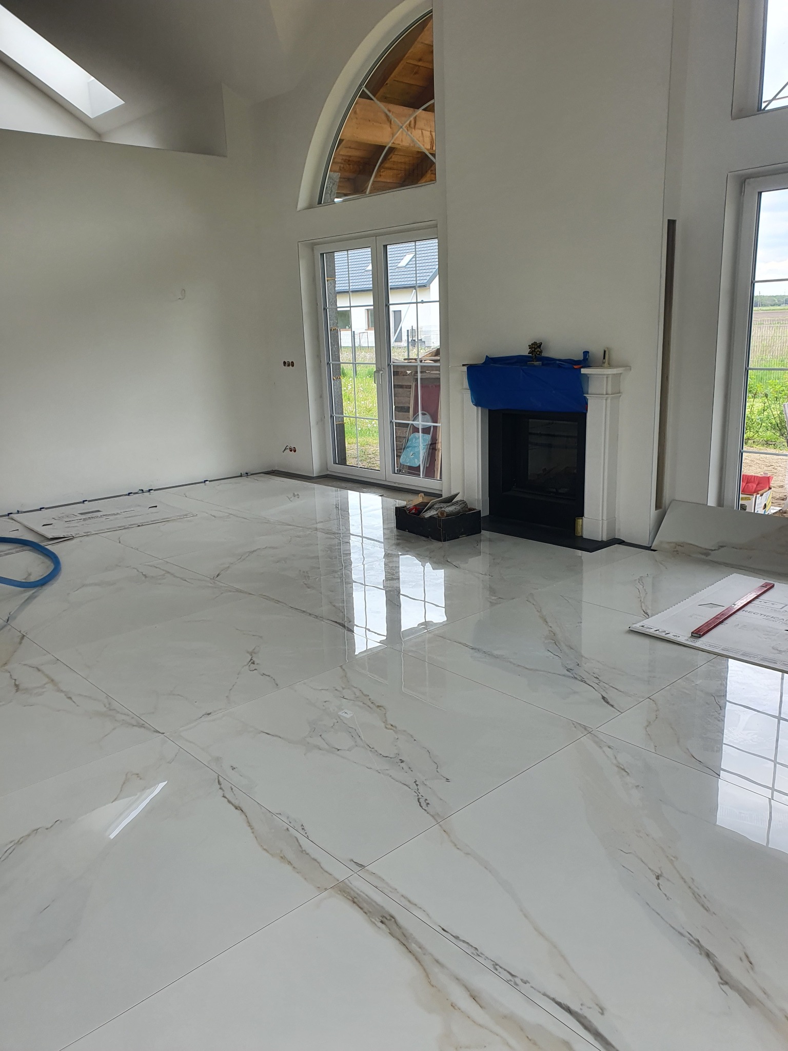 Błyszczące, białe płytki podłogowe z marmurkowym wzorem, ułożone w przestronnym salonie z kominkiem i dużymi oknami. Widoczne narzędzia i akcesoria montażowe.