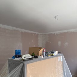 GrandFinish(RuslanYaniv) - Pokój w trakcie malowania: ściany pokryte gruntem, zabezpieczone meble, narzędzia malarskie na wierzchu. Widoczne listwy dekoracyjne pod sufitem i puszka z farbą.