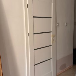 GrandFinish(RuslanYaniv) - Białe drzwi wewnętrzne z poziomymi, ciemnymi paskami, zamaskowany otwór na klamkę, ściana po malowaniu, widoczne gniazdka elektryczne, płytki podłogowe.