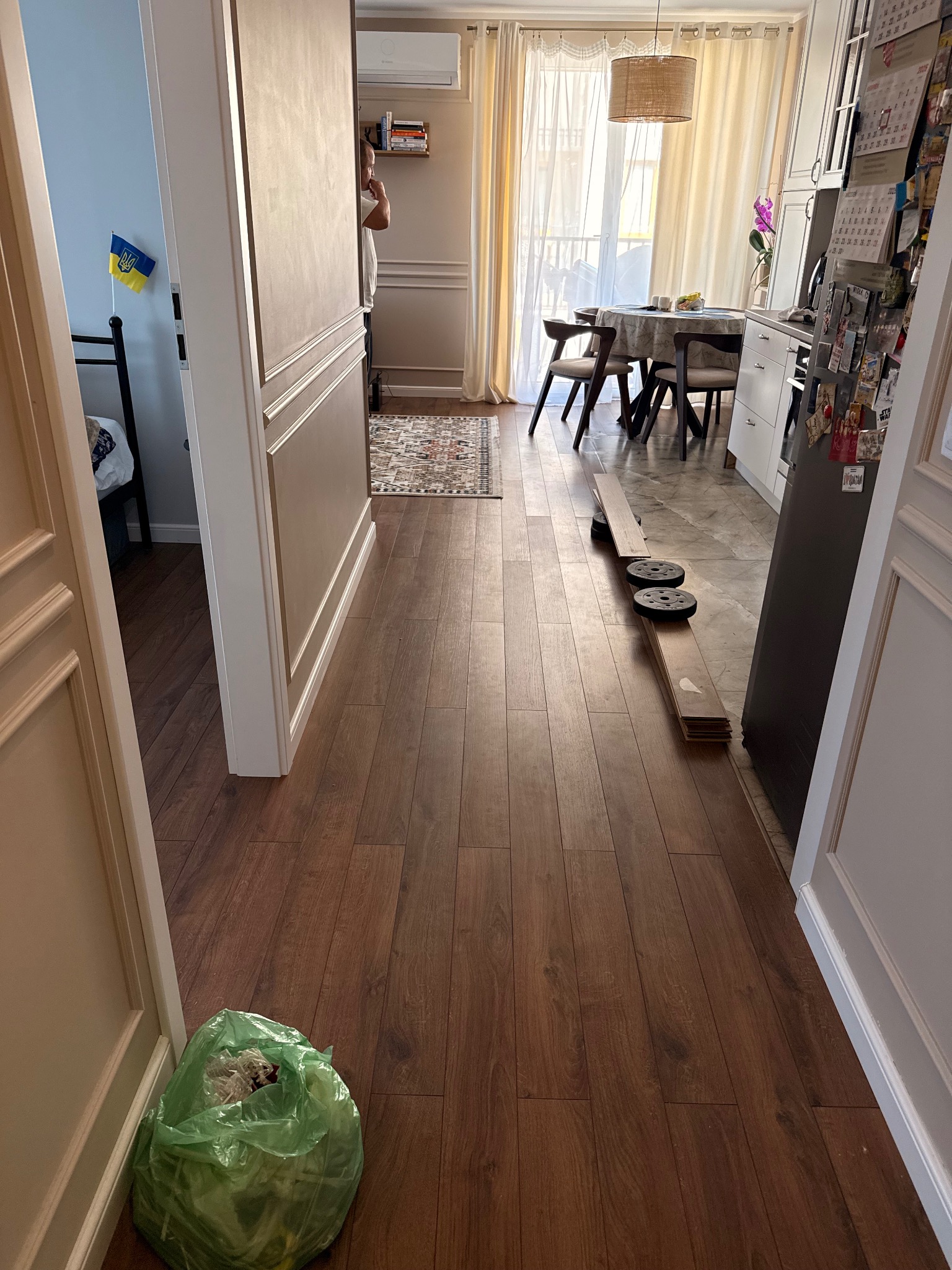 Ułożone panele podłogowe w mieszkaniu w Warszawie, widok z perspektywy wejścia do pokoju. Na podłodze leżą panele z obciążeniem i worek ze śmieciami.