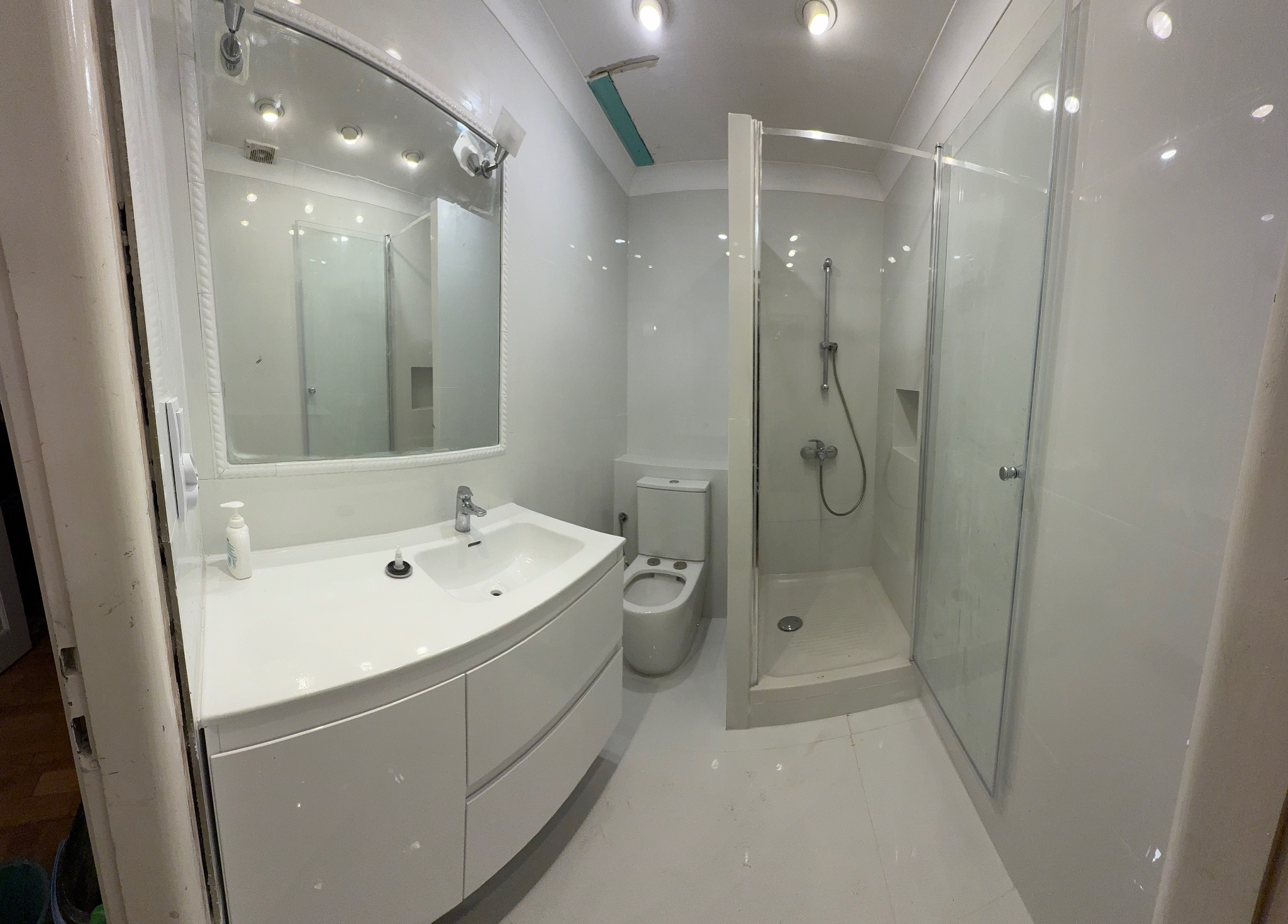 Jasna łazienka po remoncie: białe płytki, kabina prysznicowa, umywalka z szafką i duże lustro. W tle toaleta i fragment uszkodzonego sufitu.