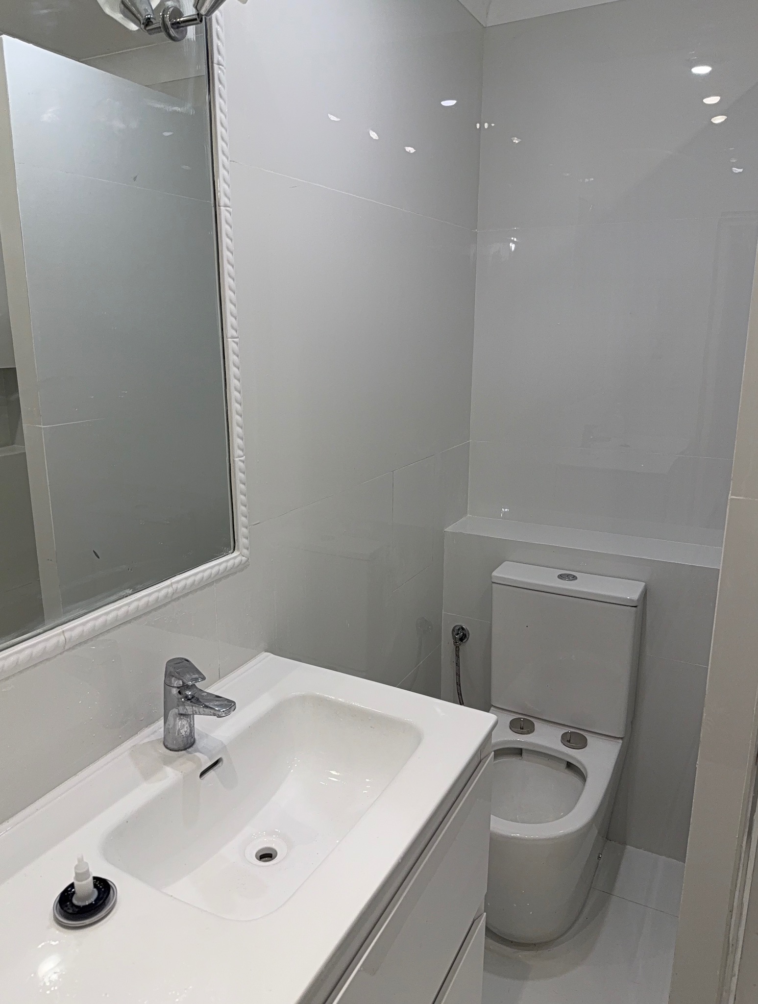 Biała, wykafelkowana łazienka po remoncie: umywalka z szafką, toaleta i lustro z ozdobną ramą. Widoczny fragment brodzika. Minimalistyczny design, czystość i porządek.