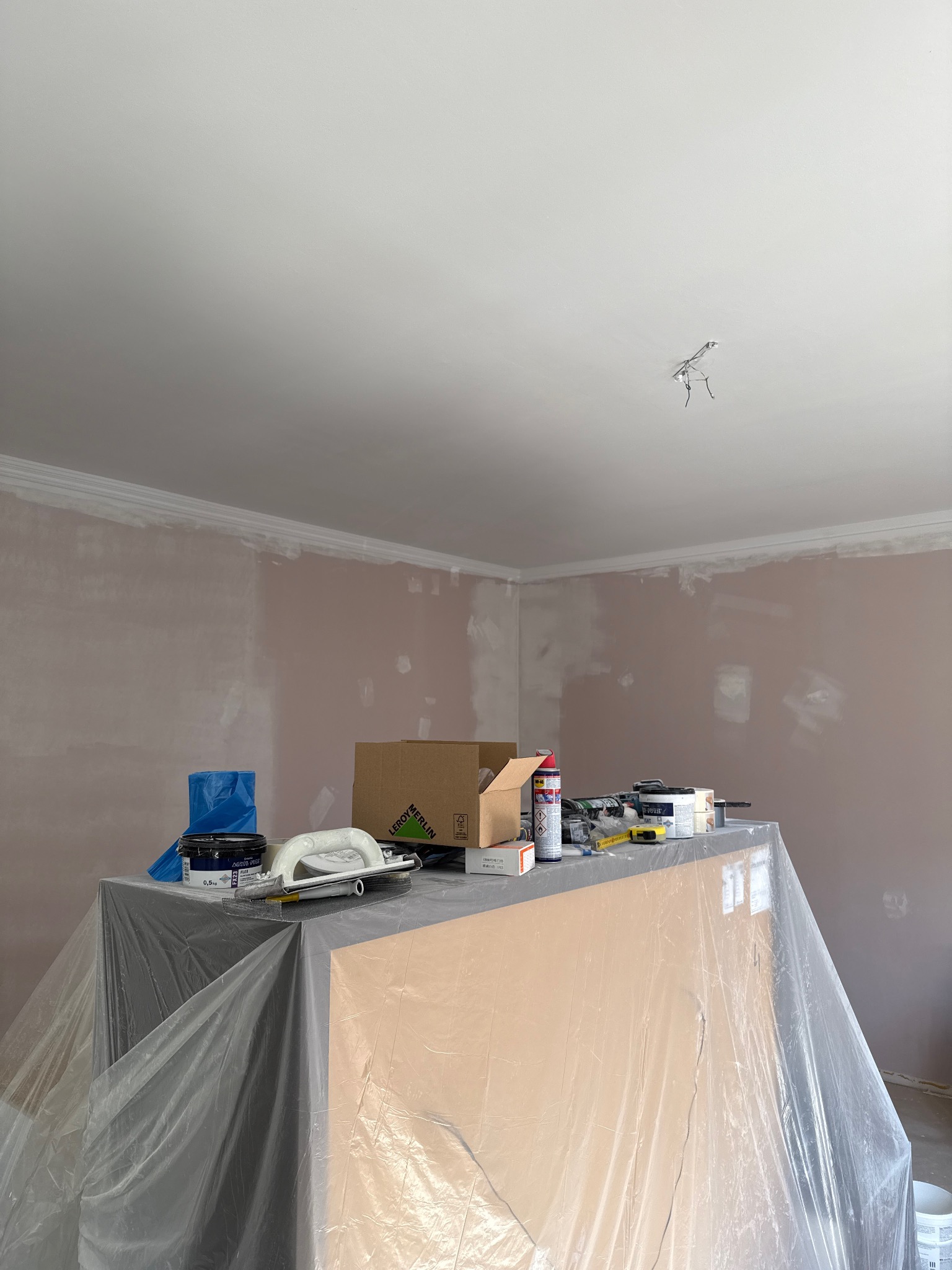 Pokój w trakcie malowania: ściany pokryte gruntem, zabezpieczone meble, narzędzia malarskie na wierzchu. Widoczne listwy dekoracyjne pod sufitem i puszka z farbą.