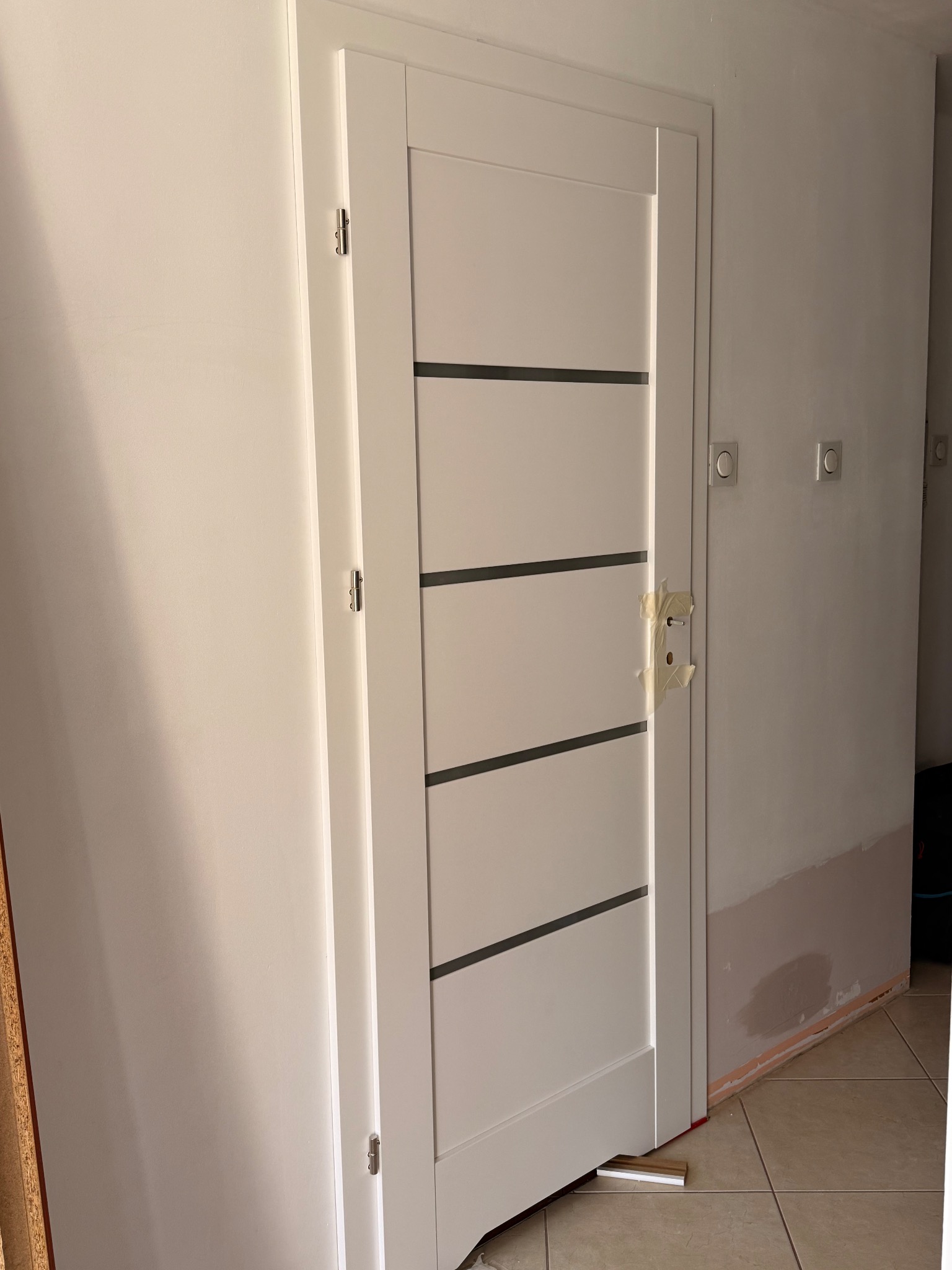 Białe drzwi wewnętrzne z poziomymi, ciemnymi paskami, zamaskowany otwór na klamkę, ściana po malowaniu, widoczne gniazdka elektryczne, płytki podłogowe.