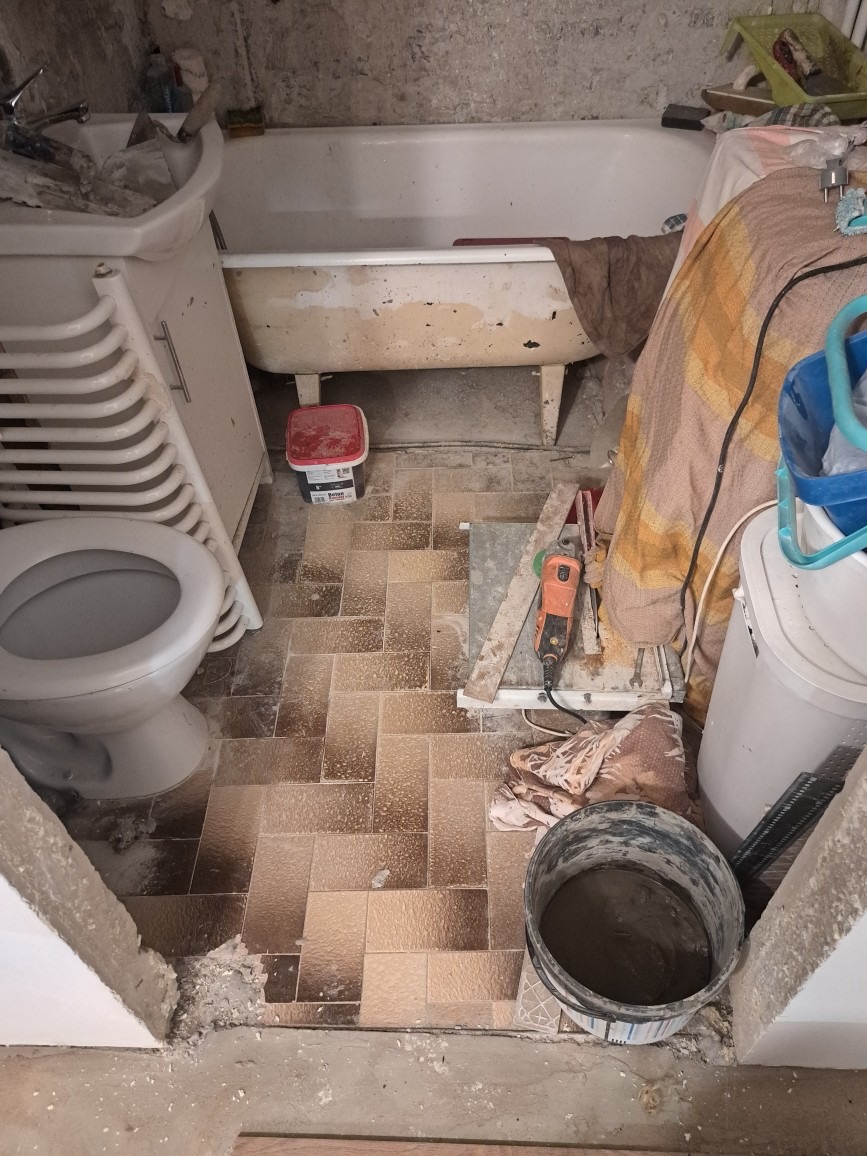 Remont łazienki w trakcie: widok na starą wannę, umywalkę, toaletę i brązowe płytki podłogowe. Obok stoi wiadro z zaprawą i szlifierka kątowa.