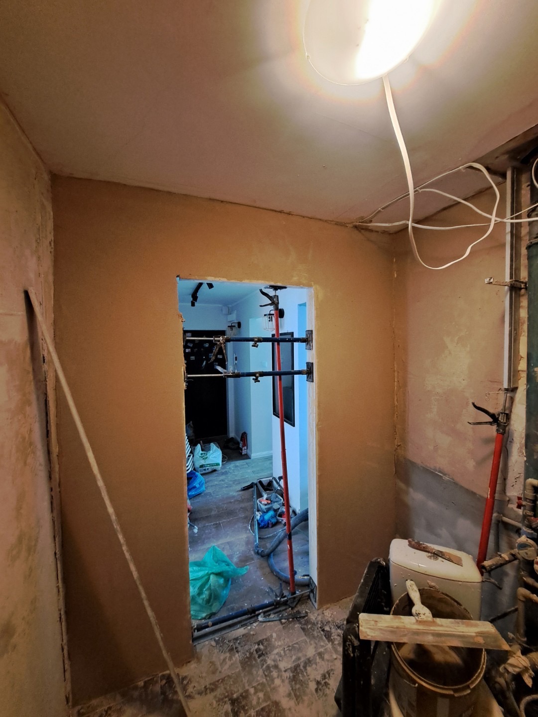 Ściana po nałożeniu tynku gipsowego, widoczne narzędzia i podpory. Przejście do innego pomieszczenia z widocznym bałaganem. Oświetlenie sufitowe.