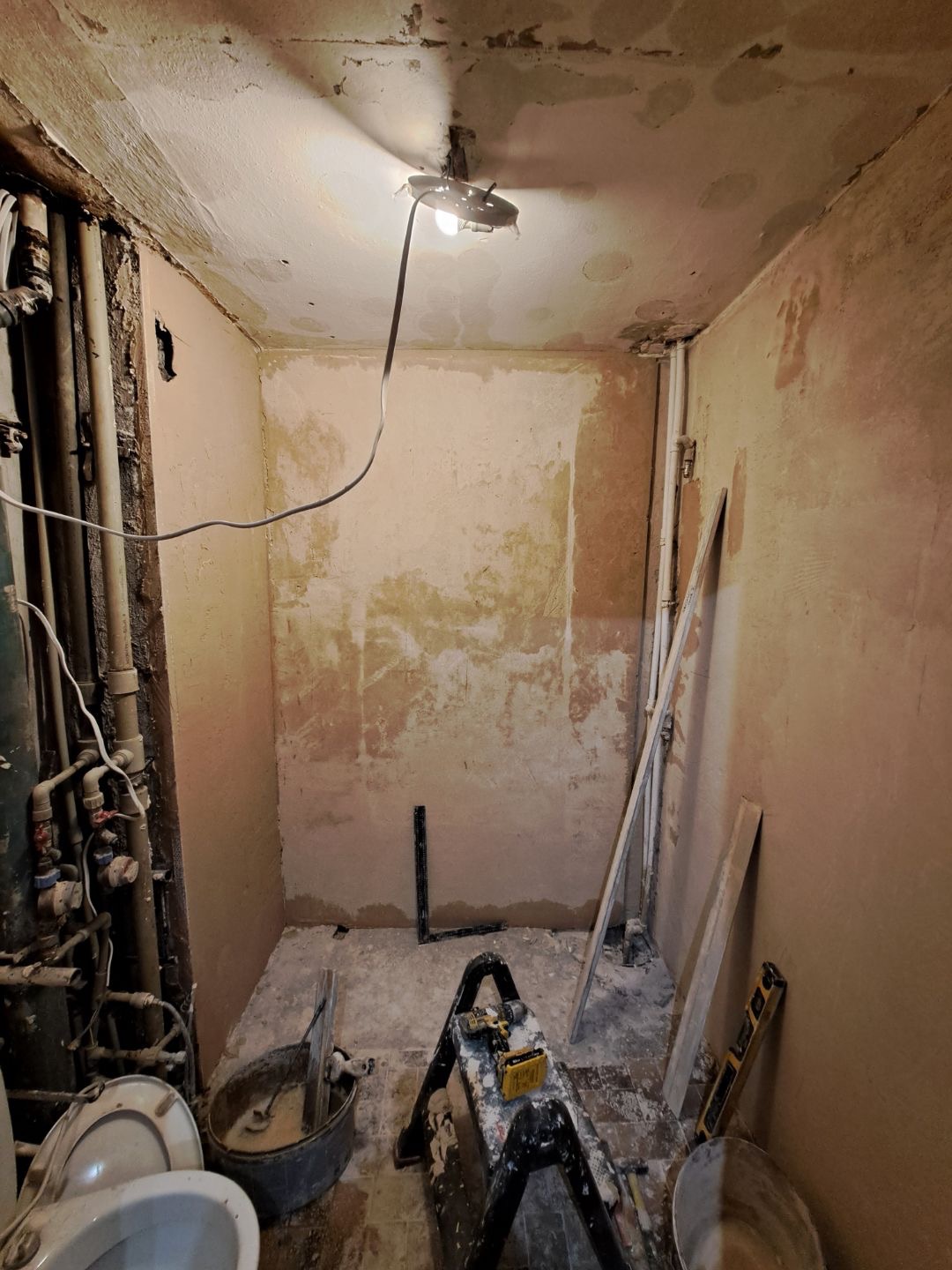 Łazienka w trakcie prac przygotowawczych: ściany po skrobaniu, widoczne instalacje, drabina malarska z narzędziami, wiadro z zaprawą. Oświetlenie robocze.