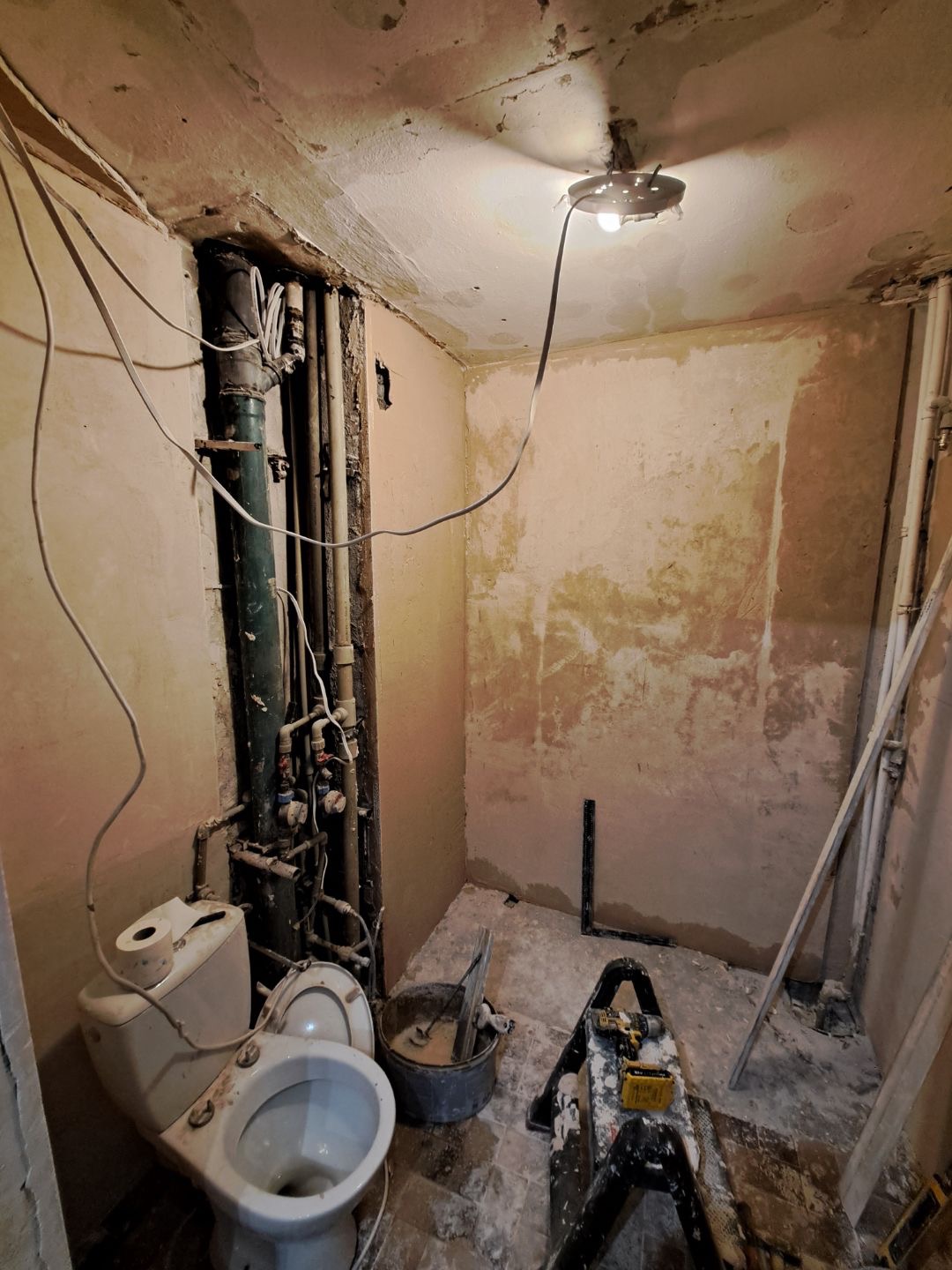 Wnętrze łazienki w trakcie remontu: widoczne rury, toaleta, narzędzia i ściany przygotowane do dalszych prac wykończeniowych. Oświetlenie robocze.