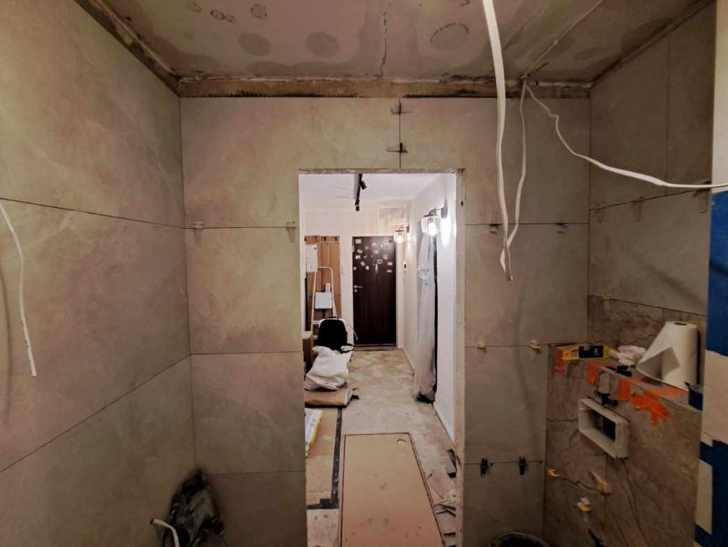 Wnętrze łazienki w trakcie prac glazurniczych. Widoczne płytki na ścianach, krzyżyki dystansowe, przewody elektryczne i narzędzia. Ujęcie z perspektywy wejścia.