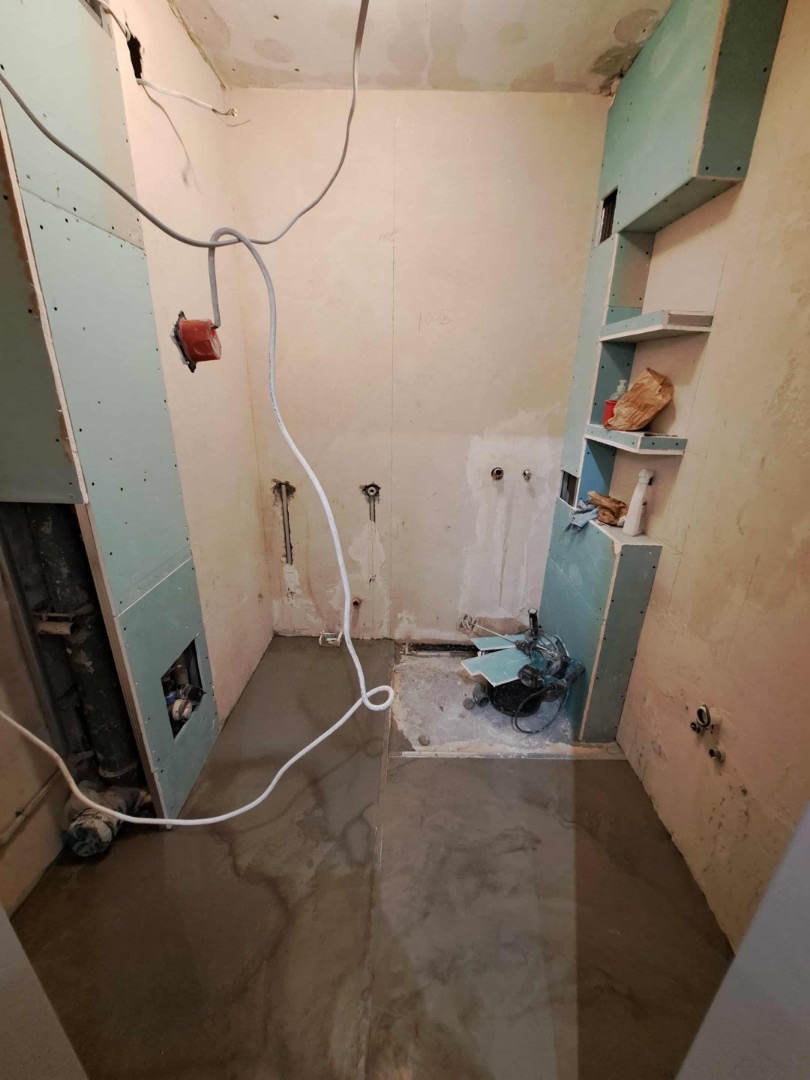 Łazienka w trakcie prac: wylewka na podłodze, płyty gipsowo-kartonowe na ścianach, widoczne przewody elektryczne i narzędzia. Ujęcie z góry, surowy stan.