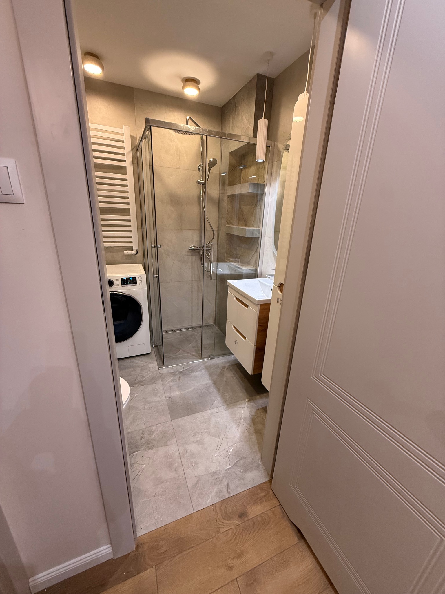 Wykończona łazienka z kabiną prysznicową, pralką i umywalką z szafką, widziana zza uchylonych drzwi. Szaro-beżowa kolorystyka, płytki imitujące marmur.