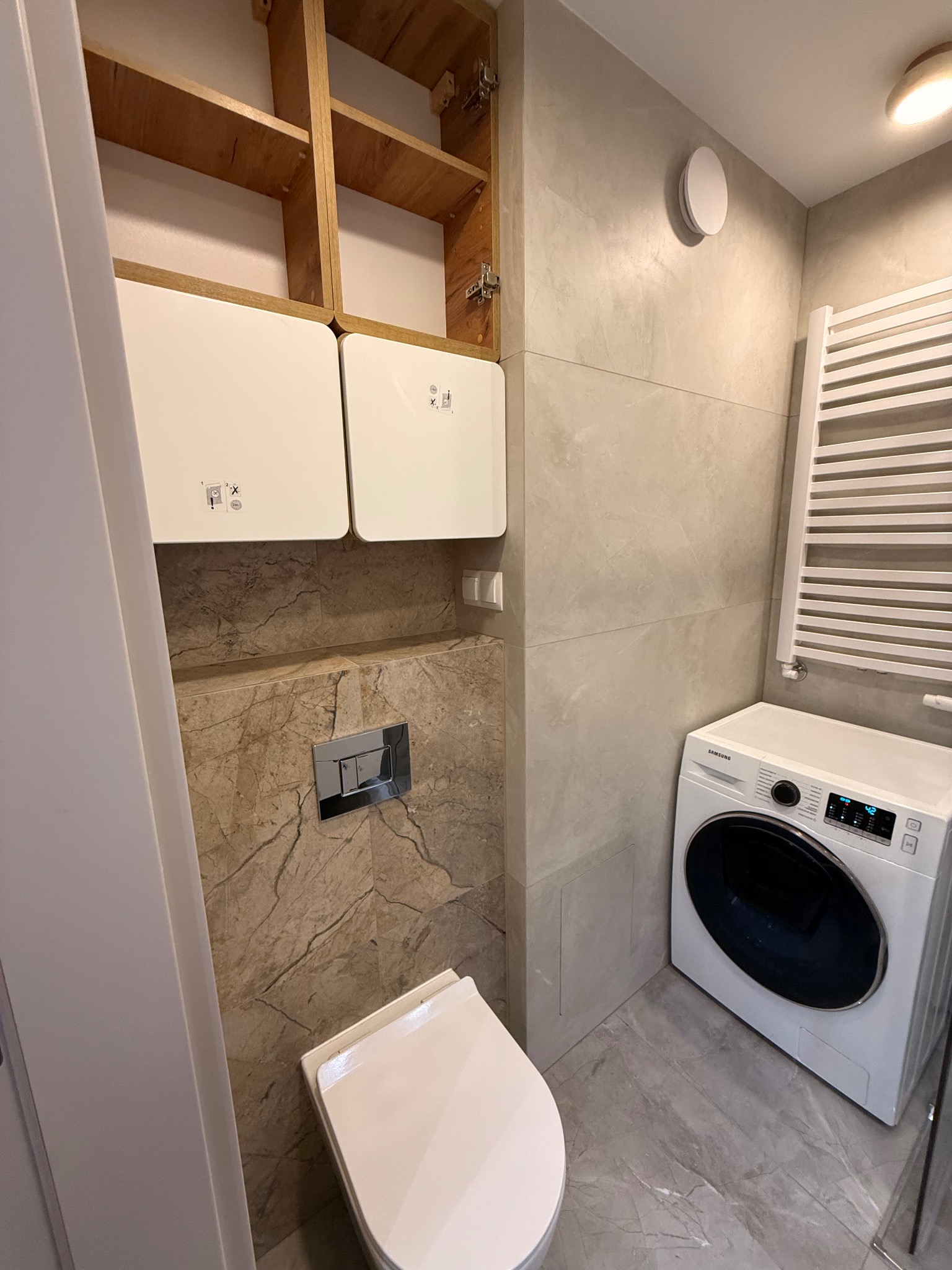 Wykończona łazienka z marmurowymi płytkami, wiszącą szafką, toaletą i pralką Samsung. Grzejnik drabinkowy na ścianie. Nowoczesny design.