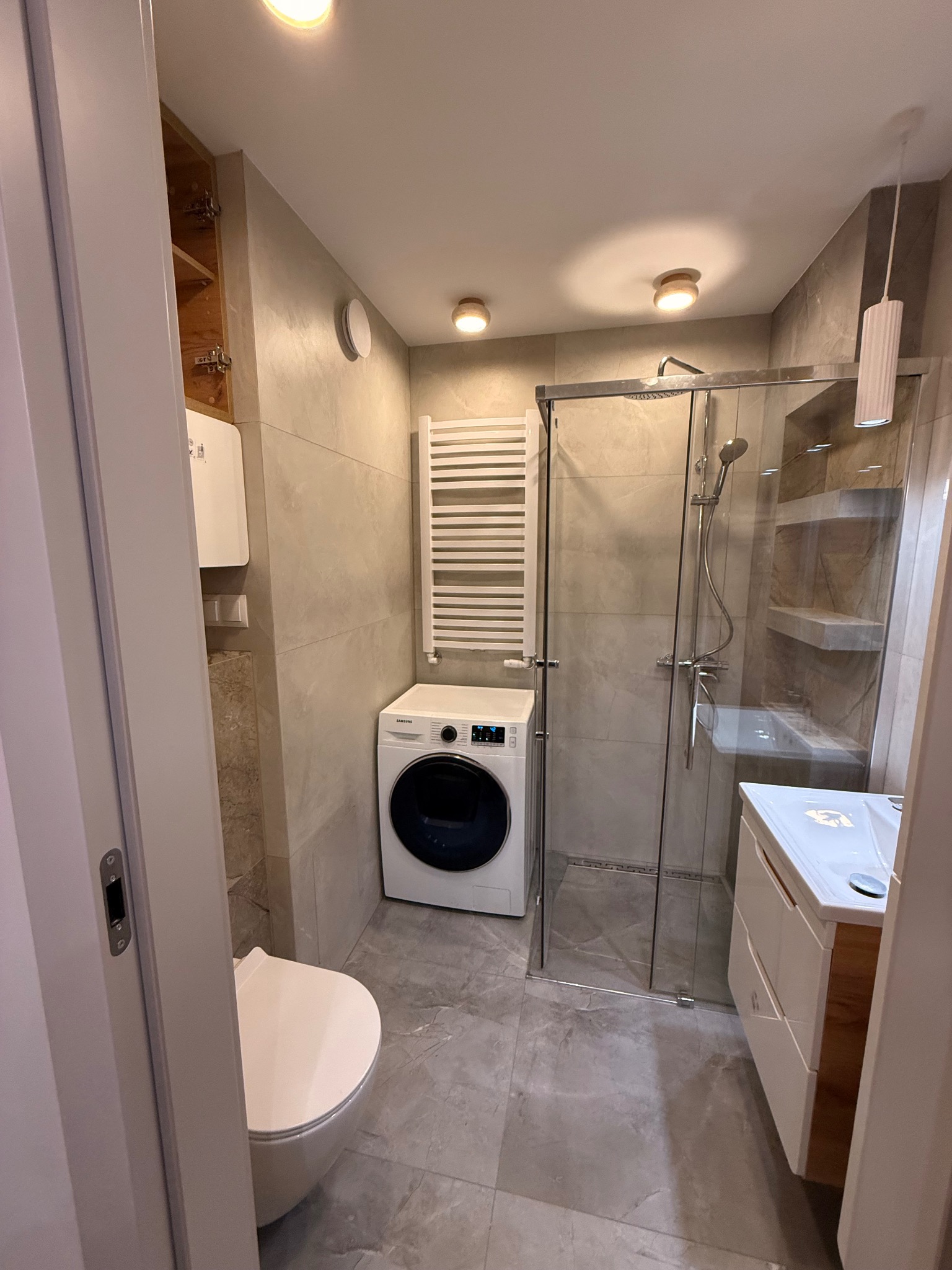 Nowoczesna łazienka w szarościach: kabina prysznicowa ze szklanymi drzwiami, umywalka z szafką, pralka i grzejnik drabinkowy. Minimalistyczne oświetlenie.