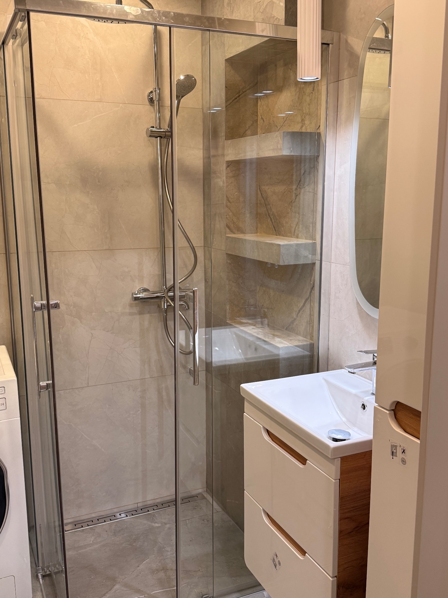 Nowoczesna kabina prysznicowa ze szklanymi drzwiami, umywalka z szafką w jasnej łazience z beżowymi płytkami. Widoczny odpływ liniowy.