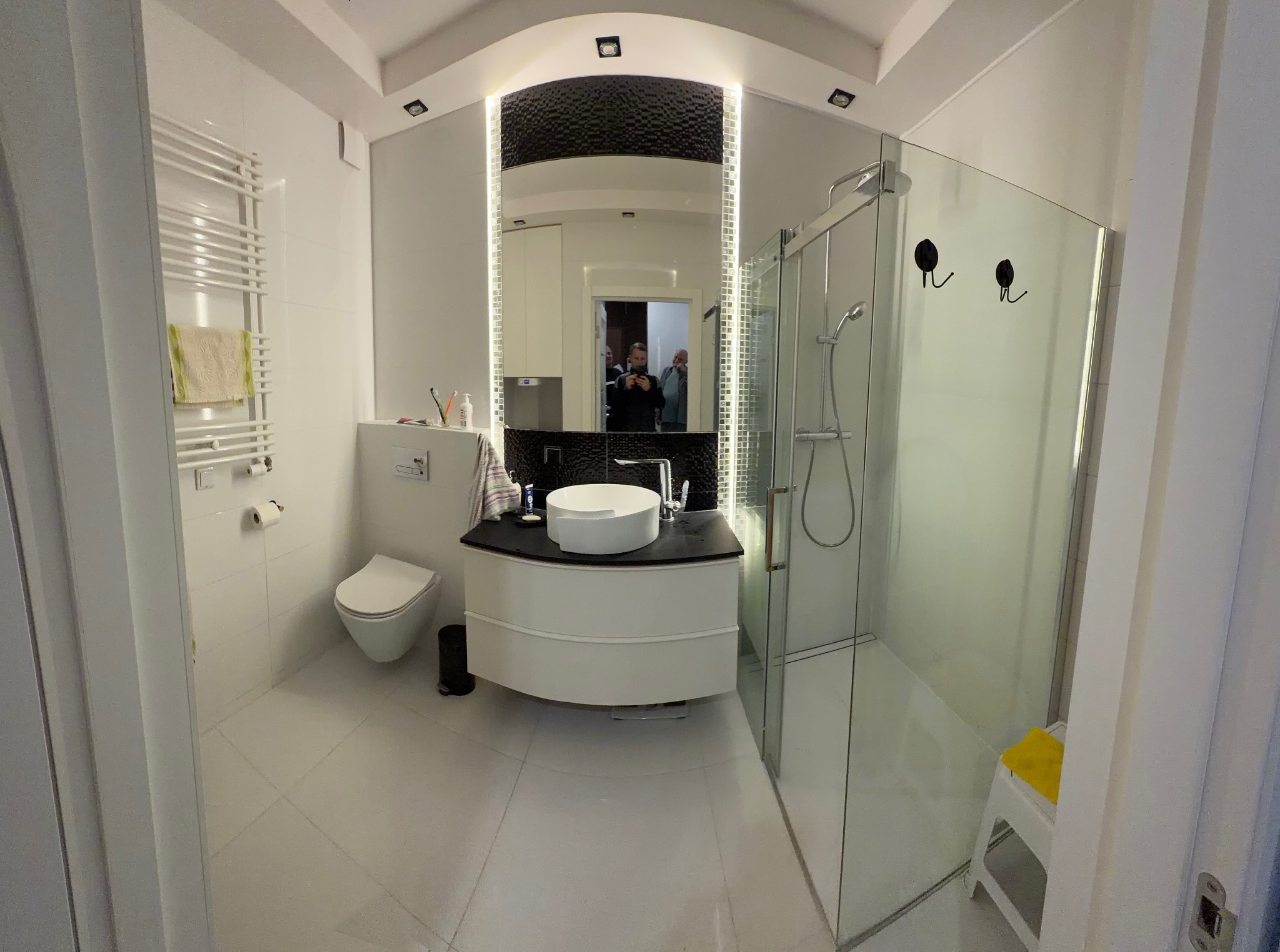 Nowoczesna łazienka z kabiną prysznicową, umywalką na szafce i podświetlanym lustrem. Białe płytki, grzejnik drabinkowy. Widoczne akcesoria łazienkowe.