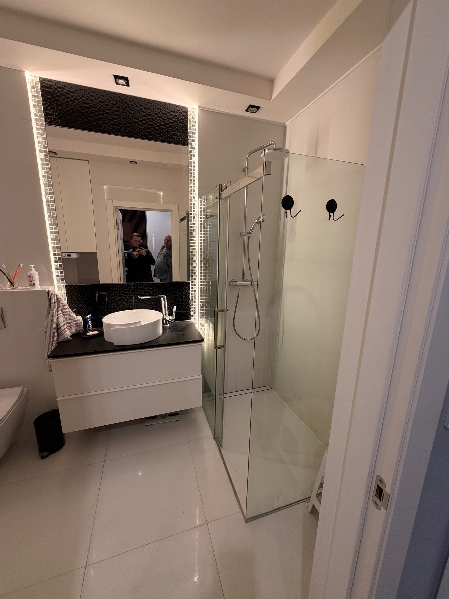 Nowoczesna łazienka z kabiną prysznicową typu walk-in, białą umywalką nablatową i lustrem z podświetleniem LED. Widoczne czarne akcenty i jasne płytki podłogowe.