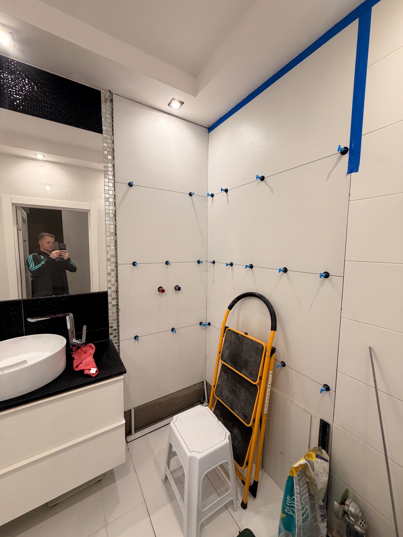 Białe płytki w łazience, poziomowane niebieskimi klipsami i sznurkami. W lustrze odbija się osoba robiąca zdjęcie. Widoczna drabina, wiadro i narzędzia glazurnicze.
