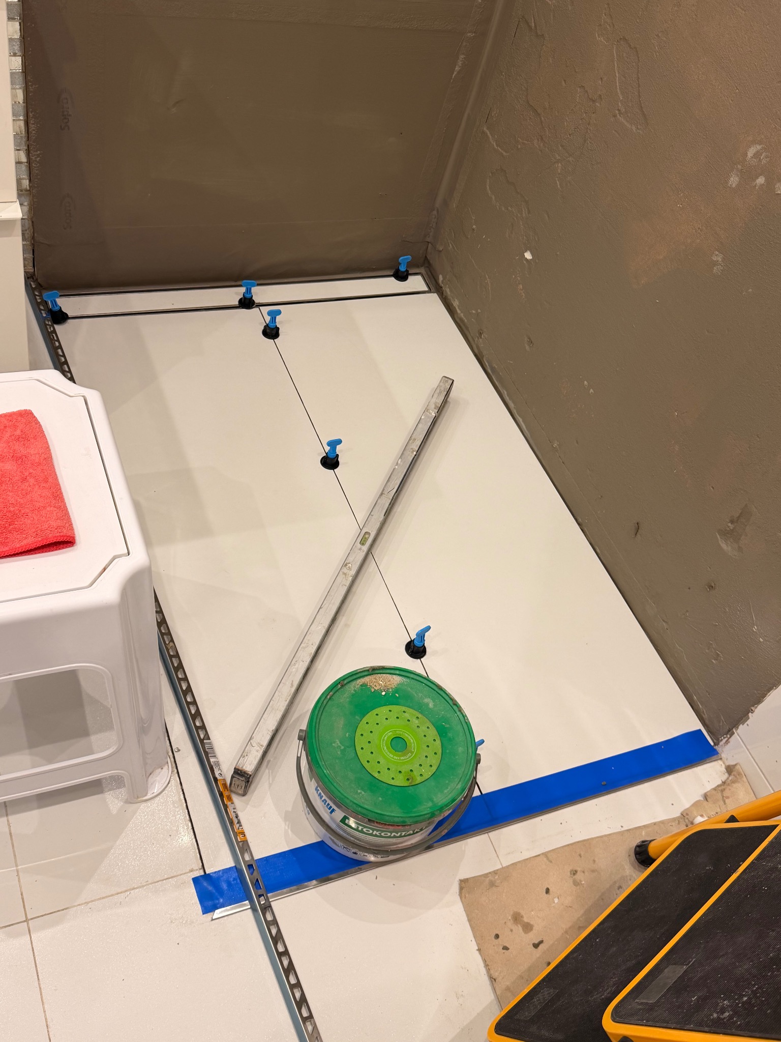 Białe płytki podłogowe w trakcie układania w łazience, z użyciem poziomicy, systemu poziomowania i wiadra z zaprawą. Widoczne zabezpieczenie ścian folią w płynie.