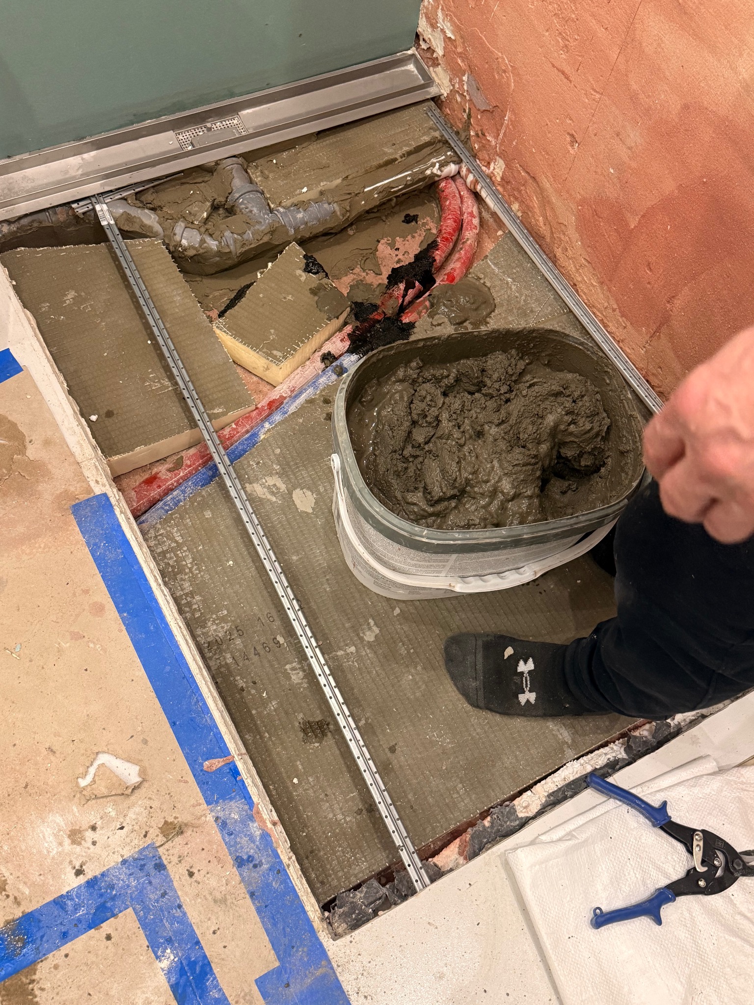 Montaż odpływu liniowego w łazience. Widoczne rury, elementy mocujące, wiadro z zaprawą i narzędzia. Prace glazurnicze w toku, przygotowanie do ułożenia płytek.