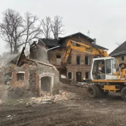 Rozbiórka ceglanego budynku w Drezdenko. Żółta koparka Liebherr w trakcie pracy, unoszący się pył i gruz. Częściowo zawalona konstrukcja.