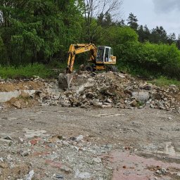 PHU Maliniak - Żółta koparka na gruzowisku po rozbiórce budynku w otoczeniu drzew, pochmurny dzień. Widoczne fragmenty fundamentów i cegieł.